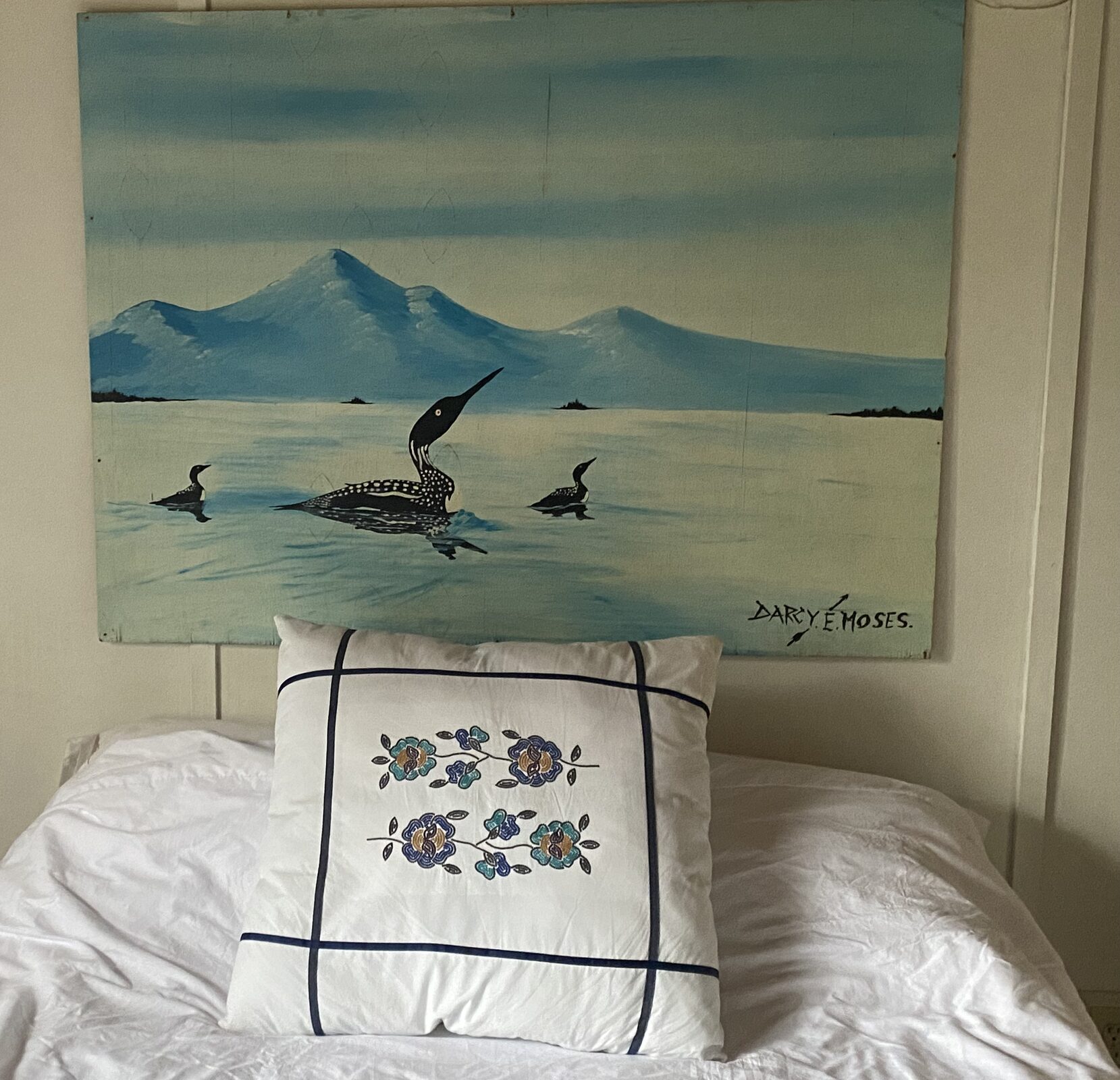 White embroidered pillow on a bed, a painting of loons on the wall behind.
